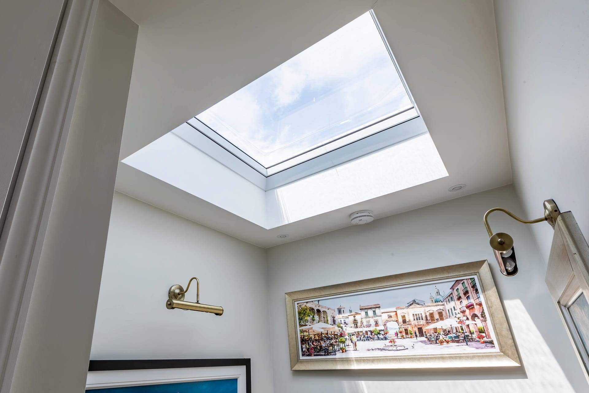 Interior wall beneath a skylight window with sunlight streaming in, highlighting framed artwork and brass wall lights.