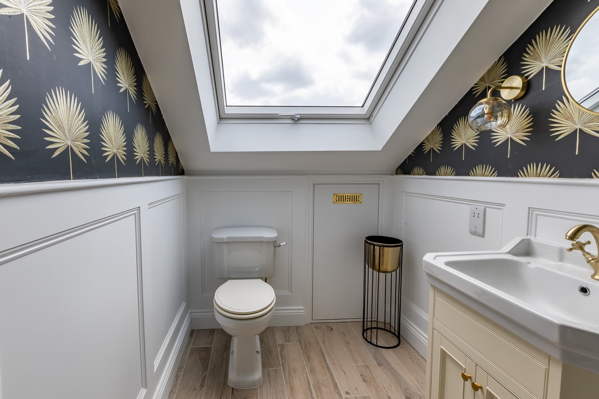 Small loft bathroom with a sloped ceiling, skylight window, white panelled walls, and black wallpaper with gold leaf pattern above.