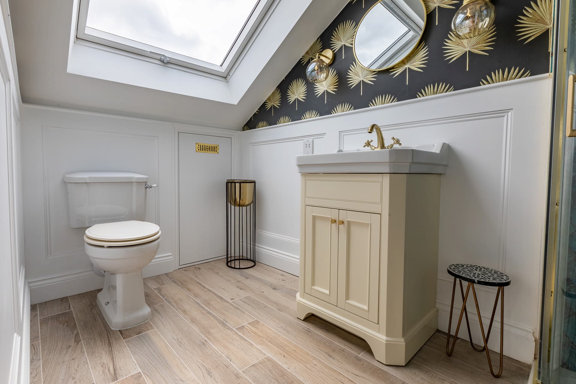 Compact loft bathroom featuring a toilet, cream vanity sink with gold fixtures, patterned black-and-gold wallpaper, and a skylight.