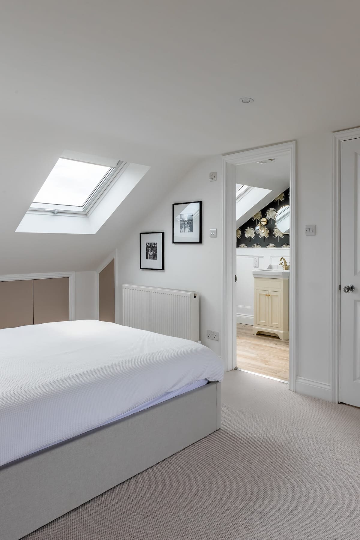 Minimalist loft bedroom with a large bed, white walls, skylight window, framed photos, and view into adjoining bathroom.