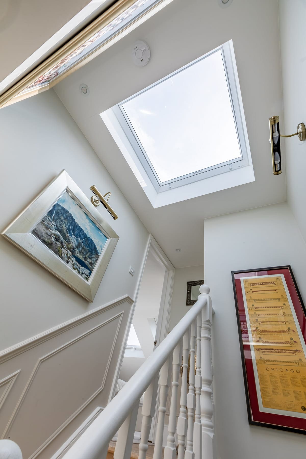 View up a staircase with white bannisters leading to a landing, illuminated by a large skylight window above and framed wall art.