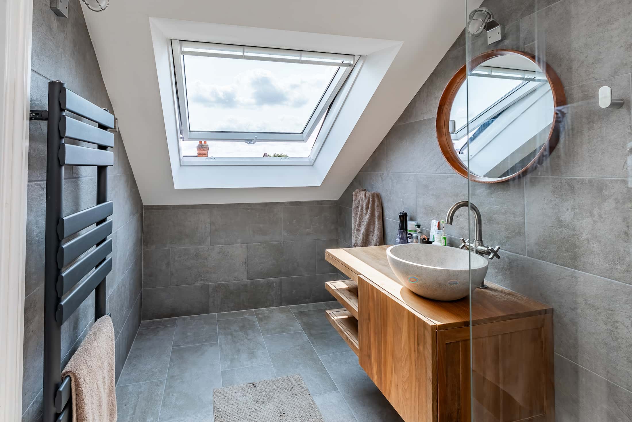 Modern loft bathroom with grey tiled walls, skylight window, wooden vanity with bowl sink, round mirror, and heated towel rail.