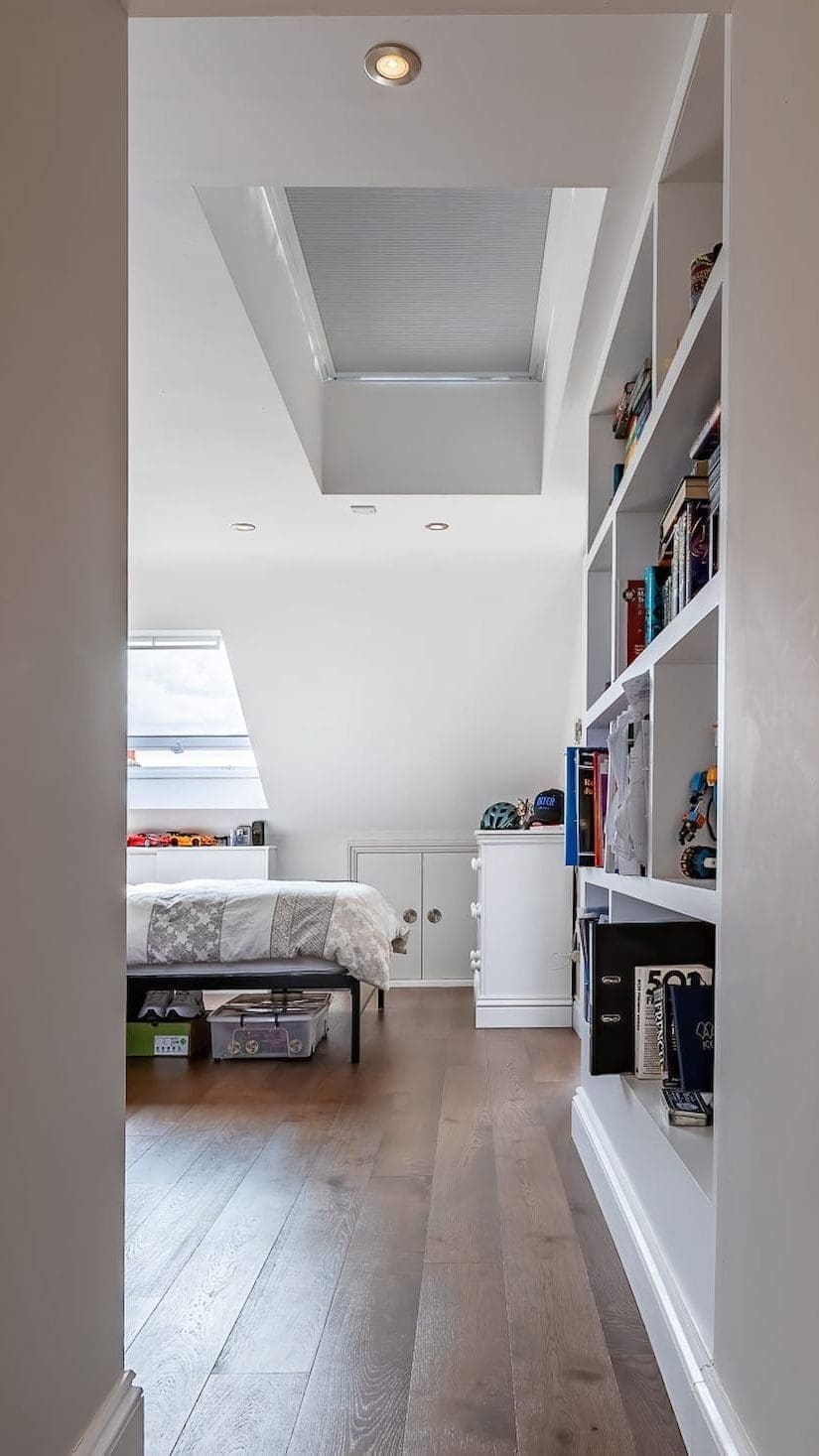 View through a doorway into a loft bedroom with wooden flooring, skylight window, bed, and built-in shelving along the wall.