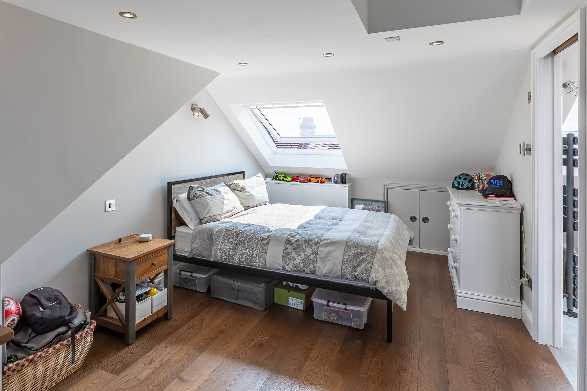 Bright loft bedroom with sloped ceiling, skylight window above the bed, wooden flooring, bedside table, and storage drawers with personal items.