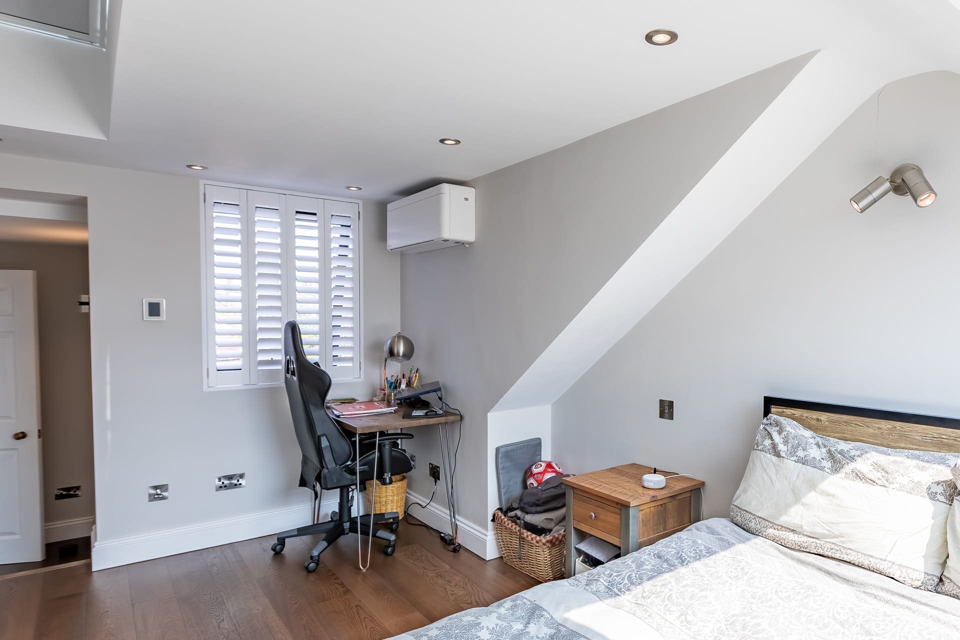 Loft bedroom with sloped ceiling featuring a bed, wooden bedside table, small desk with chair by a shuttered window, and wooden flooring.