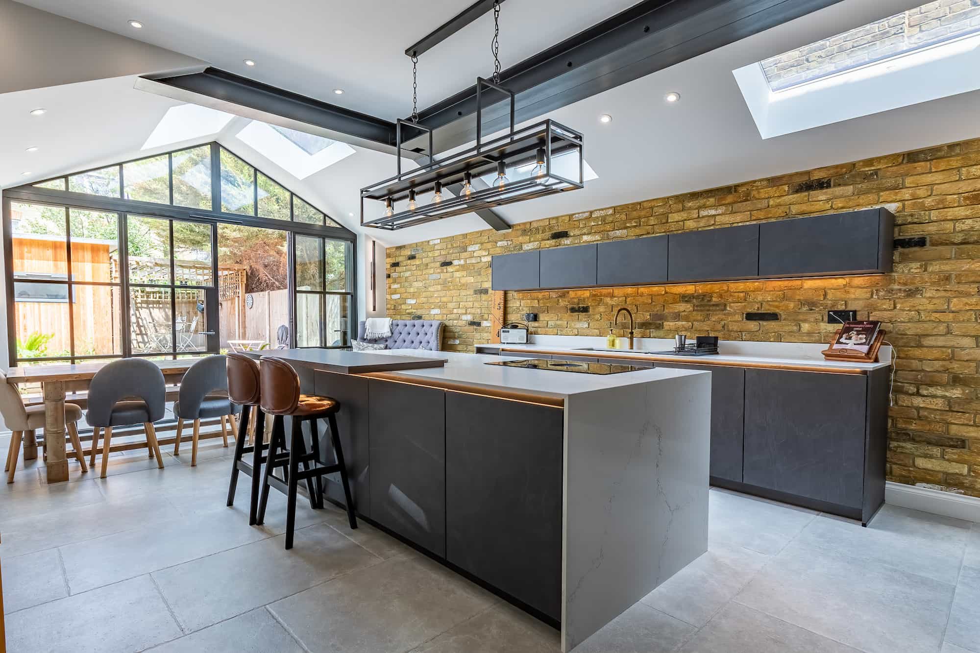 Contemporary open-plan kitchen featuring a large island with seating, exposed brick wall, skylights, pendant lighting, and dining area with glass doors leading outside.