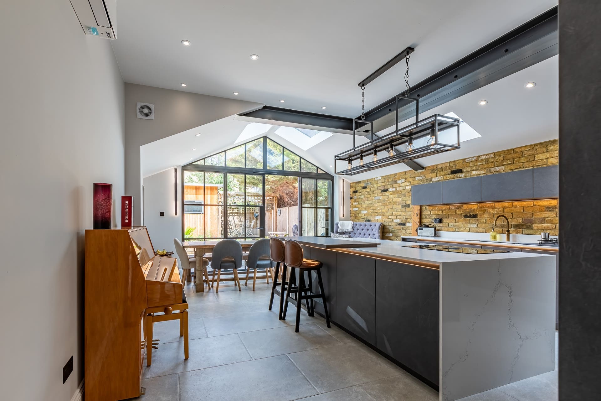 Modern open-plan kitchen with large island, pendant lighting, exposed brick wall, skylights, dining table, and floor-to-ceiling glass doors opening to a garden.