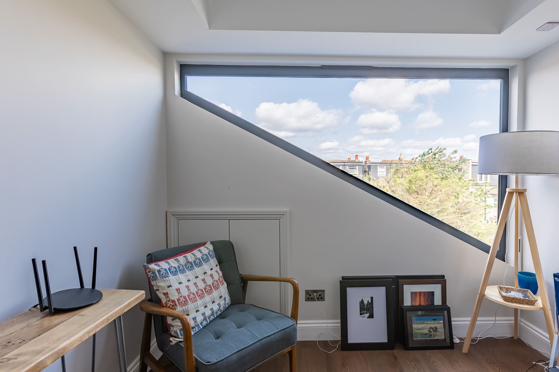 Cozy corner with angled feature window, armchair, floor lamp, wooden desk, and framed artwork leaning against the wall.
