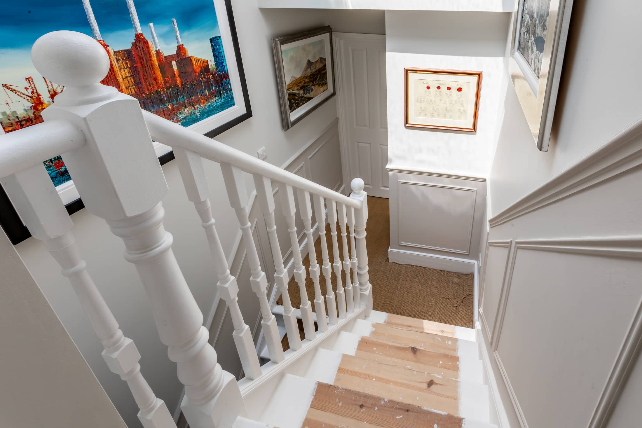 White staircase with wooden steps and decorative spindles, leading down to a hallway with framed artwork and wall panelling.