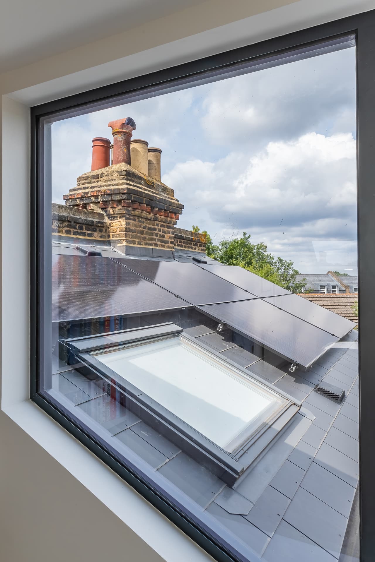 View through a loft window showing rooftops with solar panels, brick chimneys, and trees under a cloudy sky.